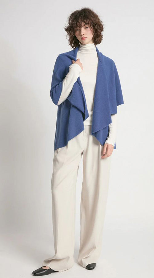 Person wearing a blue shawl over a white turtleneck and beige pants on a white background
